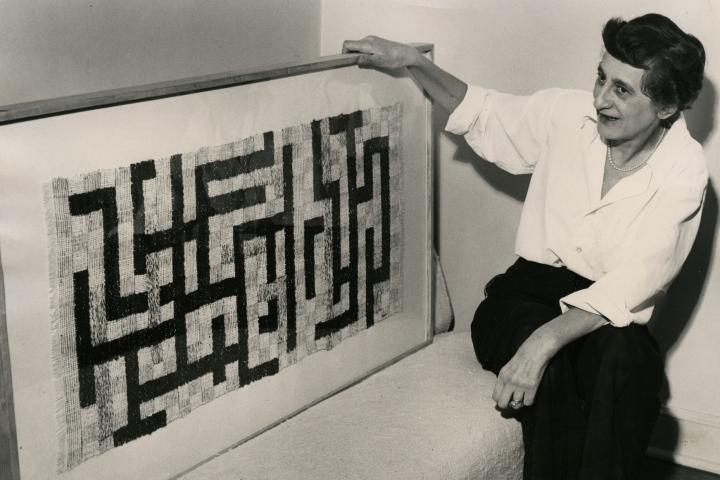 Foto einer älteren Frau, die neben einem geometrischen Textilkunstwerk sitzt.